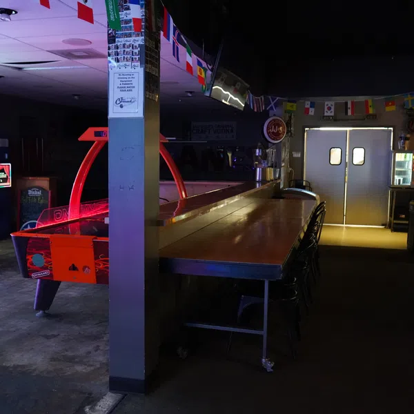 Dimly lit arcade room with air hockey table, arcade games, bar counter, and international flags overhead.
