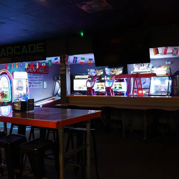 Interior of a dimly lit arcade with neon-lit game machines and high tables with stools.