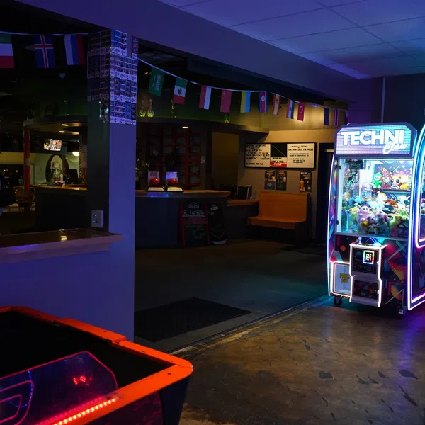 Arcade claw machine glowing with neon lights in a dimly lit recreational room with flags and a pool table.