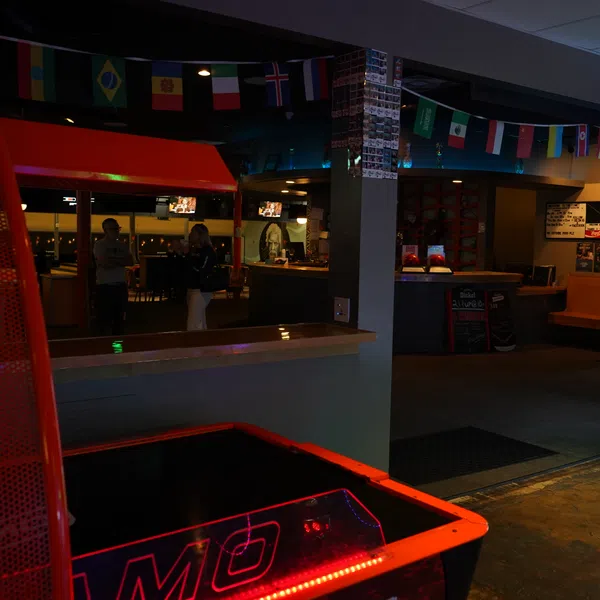 Indoor arcade with basketball game, claw machine, international flags, and people at a bar counter in the background