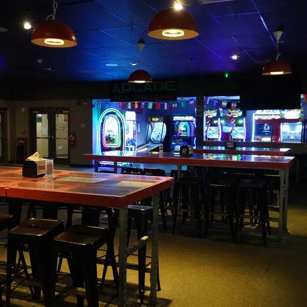 Dimly lit arcade with glowing game machines, tall tables, and stools under hanging pendant lights.