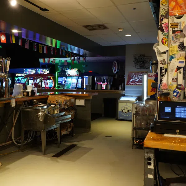 Interior of a dimly lit bar featuring a wooden counter, beer taps, arcade machines, and a cluttered cashier area.