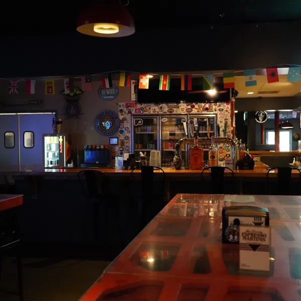 Dimly lit bar interior with international flags, stools at counter, and a neon Triple beer sign.