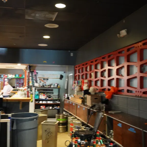 Bar interior with shelves holding bottles, kegs on floor, red geometric wall design, and staff in background.