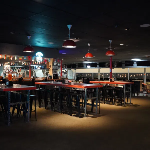 Dimly lit bowling alley with red tables, black stools, hanging lights, and a bar area decorated with international flags.