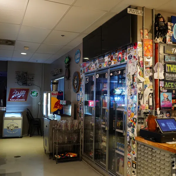 Interior view of a bar with a fridge covered in stickers, a cash register, and various drink logos on the walls.