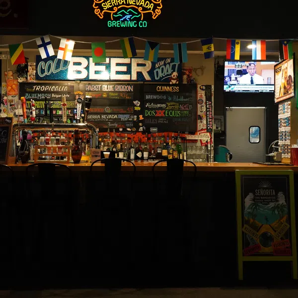 Dimly lit bar with multiple international flags, various beer signs, and black bar stools.