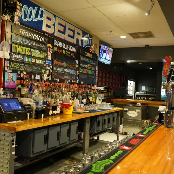 Interior of a bar with a wooden counter, liquor bottles, beer taps, and a colorful beer menu on the wall.