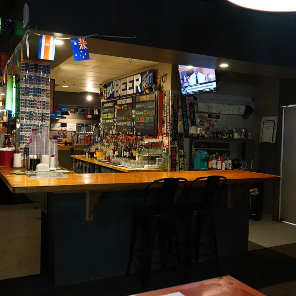 Cozy bar interior with wooden counter, bar stools, hanging flags, beverage menu, and TV screen showing news.