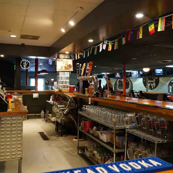 Interior view of a cozy bar with wooden counters, beer taps, international flags, and glassware on shelves.