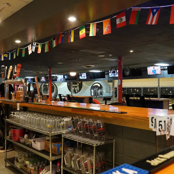Long wooden bar in a bowling alley with beer taps, shelves of glasses, and international flags above.