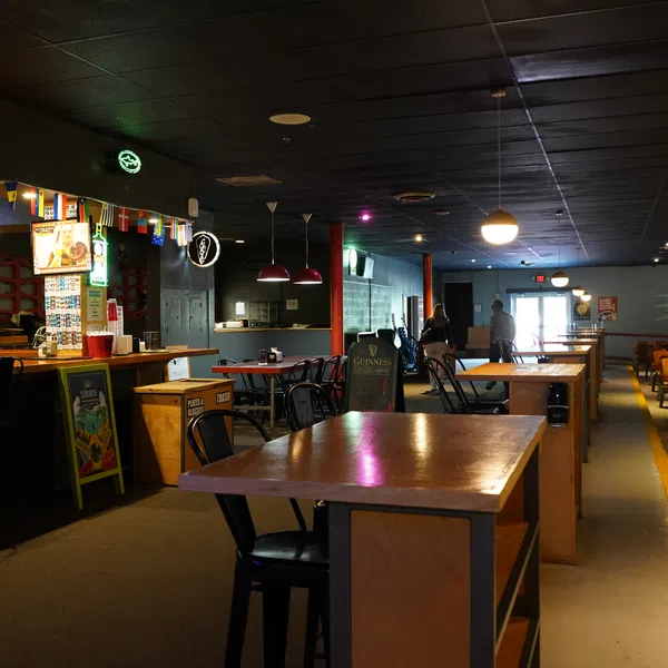 Interior of a dimly lit bar with wooden tables, chairs, neon signs, and a long bar counter with stools.