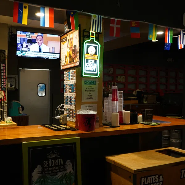 Dimly lit bar interior with wooden counter, international flags overhead, neon liquor sign, and TV screen showing news.