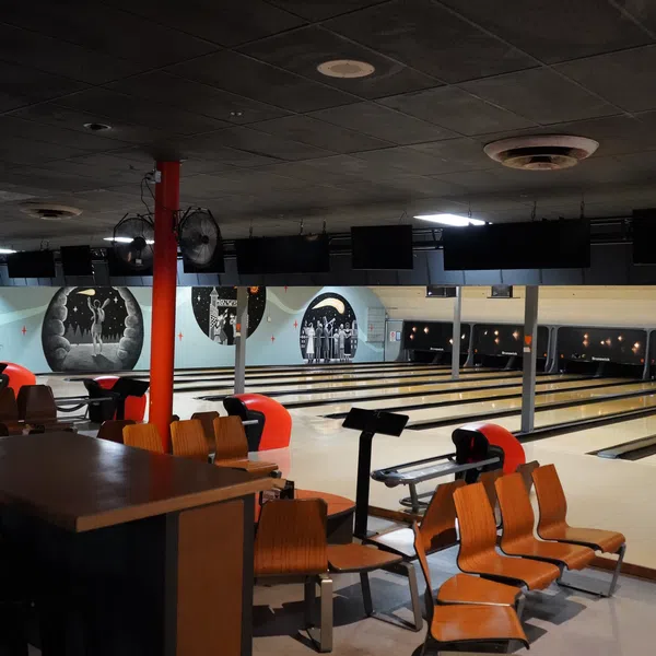 Interior view of a retro bowling alley with wooden chairs, lanes, and black and white wall murals under dim lighting.