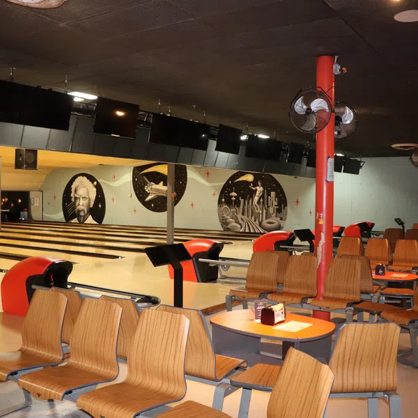 Interior of a retro-style bowling alley with wooden chairs, round tables, and mural artwork on the walls.