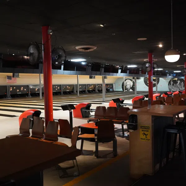 Interior view of a dimly lit bowling alley with empty lanes, seating, and tables under ceiling fans and lights.
