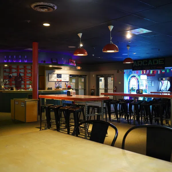 Empty arcade and bar area with high tables, stools, neon lights, and arcade machines in a dimly lit room.