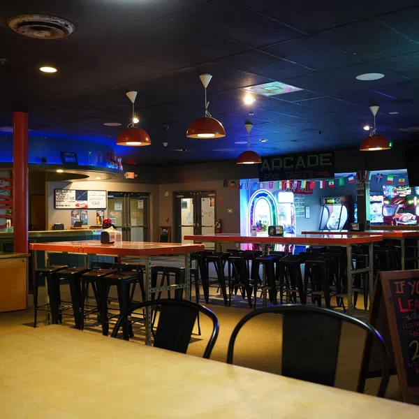 Interior of a dimly lit arcade and bar area with stools, neon lights, and hanging lamps.