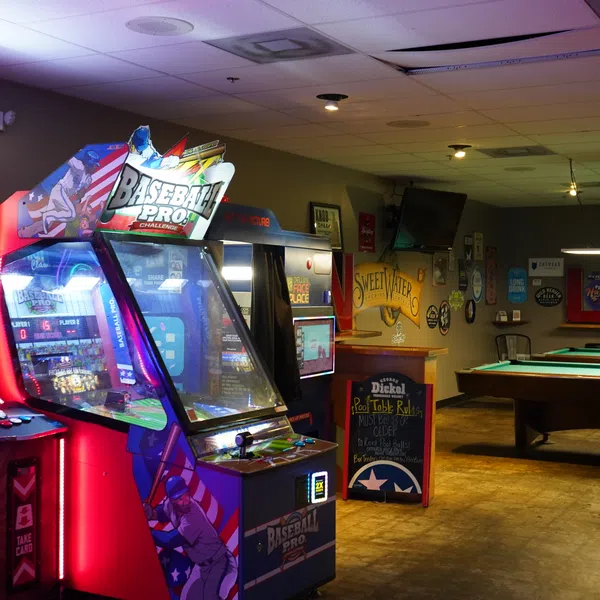Arcade room with illuminated Baseball Pro and Justice arcade machines next to a pool table in a dim bar.