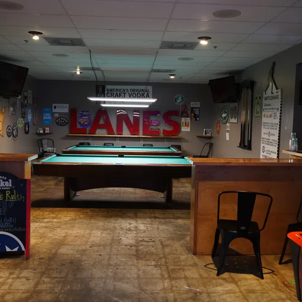Interior of a cozy pool hall with pool tables, bar area, neon signs, and casual seating in a rustic setting.