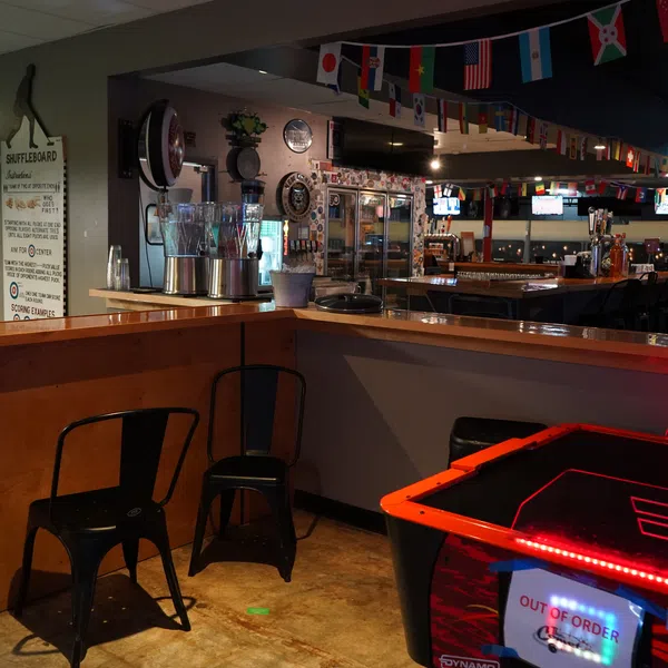 Dimly lit bar interior with shuffleboard rules, black chairs, flags, and an out-of-order arcade game at the counter.