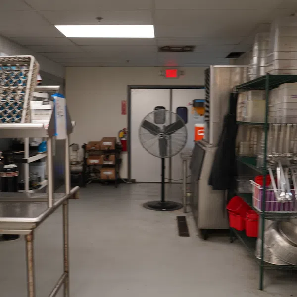 Industrial kitchen dishwashing area with metal racks, cleaning supplies, and a large standing fan.