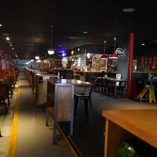 Interior view of a spacious bar with wooden chairs, tables, and a colorful illuminated bar counter under dim lighting.
