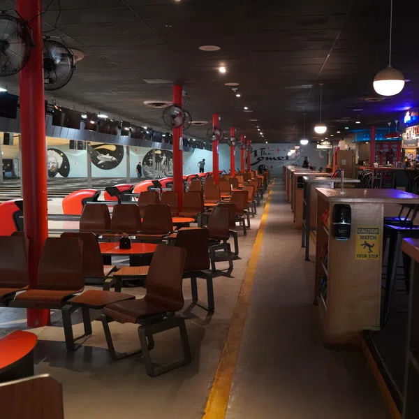 Interior view of a retro bowling alley with wooden seating, red tables, and a long bar area under dim lighting.