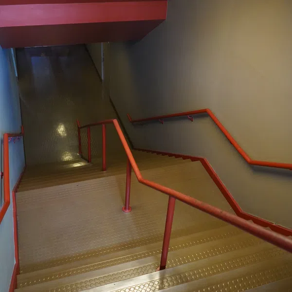Indoor staircase with textured metal steps and red handrails descending into a dimly lit corridor.