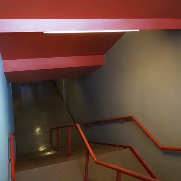 Indoor stairwell with red handrails and ceiling, gray walls, leading down to a dimly lit corridor.