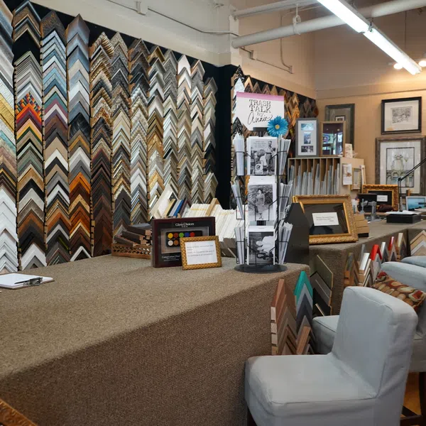 Interior of a frame shop with wall display of frame corners, workstations, and seating area with upholstered chairs.
