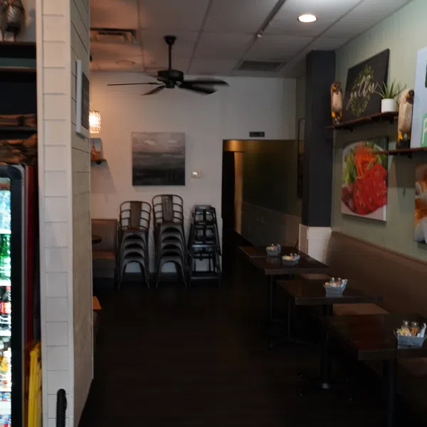 Interior of a small restaurant with stacked chairs, tables, wall art, and a refrigerator of bottled drinks.