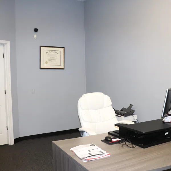 Modern office with a wooden desk, white leather chair, dual monitors, and framed certificate on light gray wall.