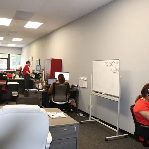 Modern office with multiple desks, computers, employees working, and a woman eating near a whiteboard on the right.
