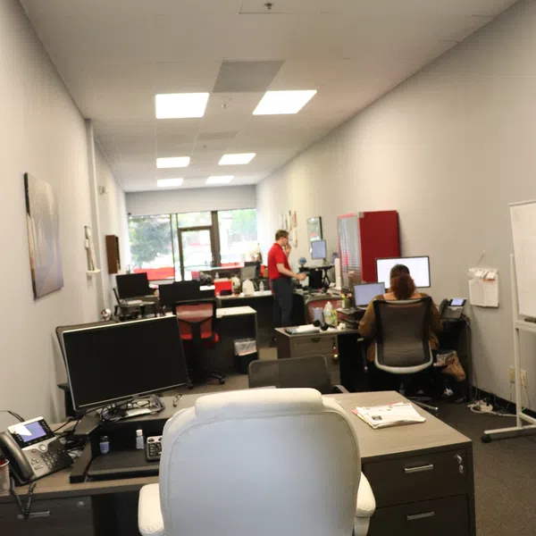 Modern office workspace with desks, computers, two people working, and overhead fluorescent lighting in a narrow room.