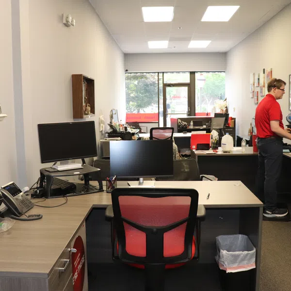 Modern office space with multiple desks, computer monitors, a standing employee, and a large window letting in daylight.