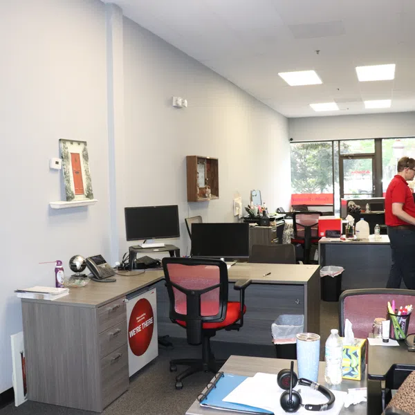 Modern office space with desks, computer monitors, office chairs, and a person working near the window.