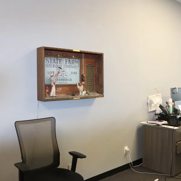 Office corner with a mesh chair, desk cluttered with items, and a vintage State Farm Insurance shadow box on the wall.