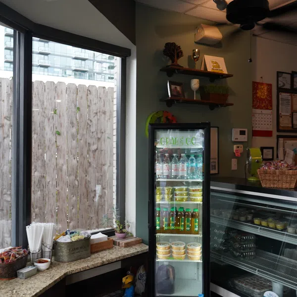 Cozy cafe corner with display fridge, condiment station, and shelves in front of large windows.