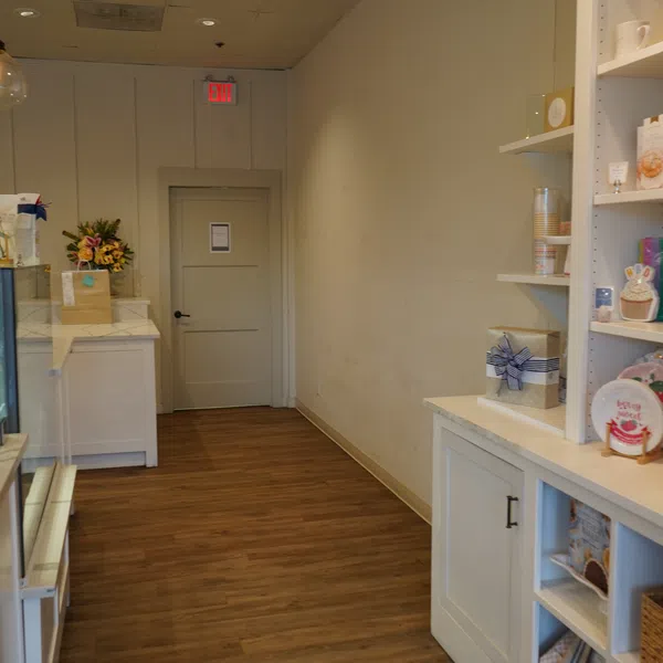 Interior view of a clean, modern bakery or cafe with wooden floor and white shelves displaying packaged goods.