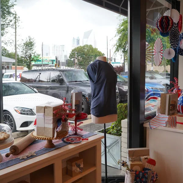 Patriotic-themed shop window with blue dress on mannequin, gift boxes, decorations, and baked goods with city street background