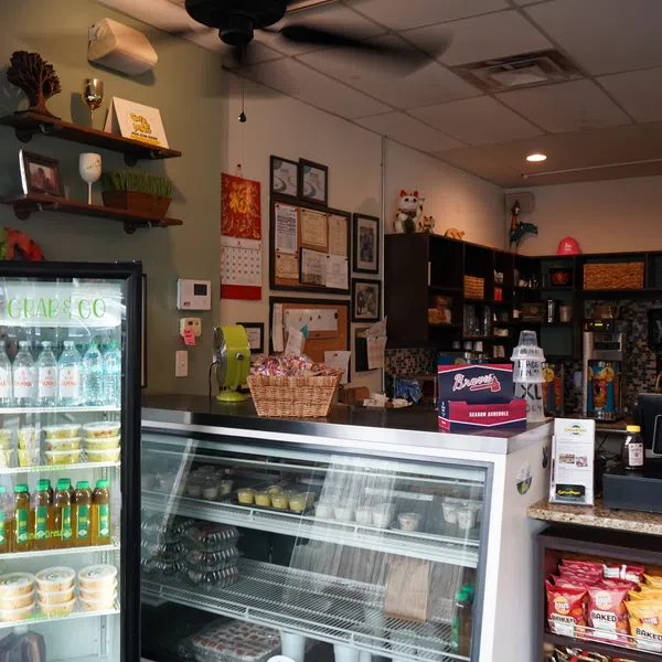 Interior of a small deli with refrigerated display cases, snacks, certificates on wall, and cash register counter.