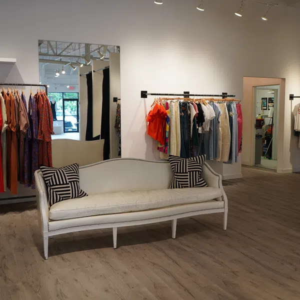Bright clothing store interior with wooden floor, racks of colorful clothes, and a white cushioned sofa with patterned pillows.