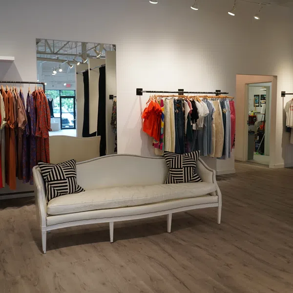 Bright boutique interior with clothing racks, a white vintage sofa, and wooden flooring under soft lighting