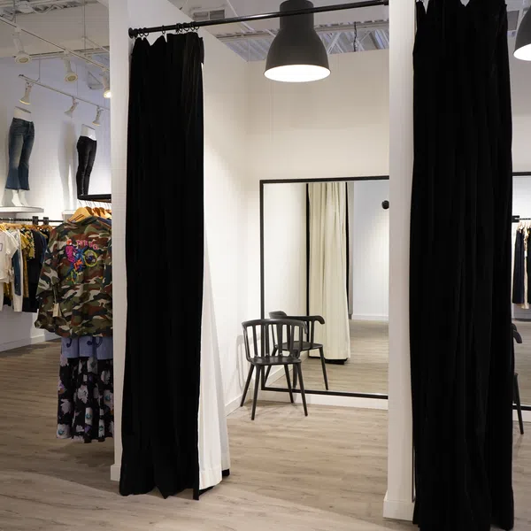 Clothing store dressing room with black curtains, wooden chair, large mirrors, and hanging apparel on racks.