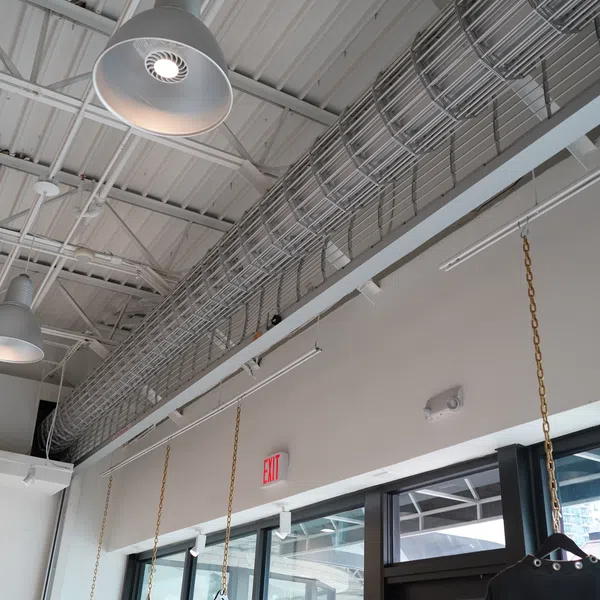 Industrial ceiling with hanging lamps, metal wiring trays, and large windows in a modern commercial space.