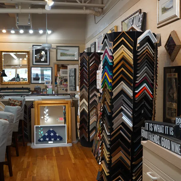 Interior of a frame shop with various frame samples, framed art, mirrors, and decorative signs on wooden floor.