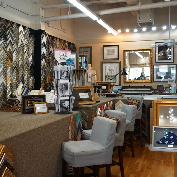 Interior of a frame shop displaying various picture frames, mirrors, and framed artworks on walls and tables.
