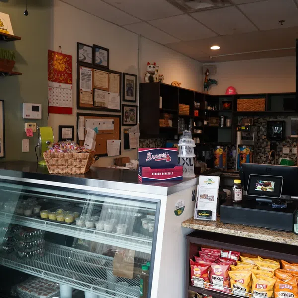 Interior of a small deli with refrigerated display, shelves of snacks, registers, and certificates on the wall.
