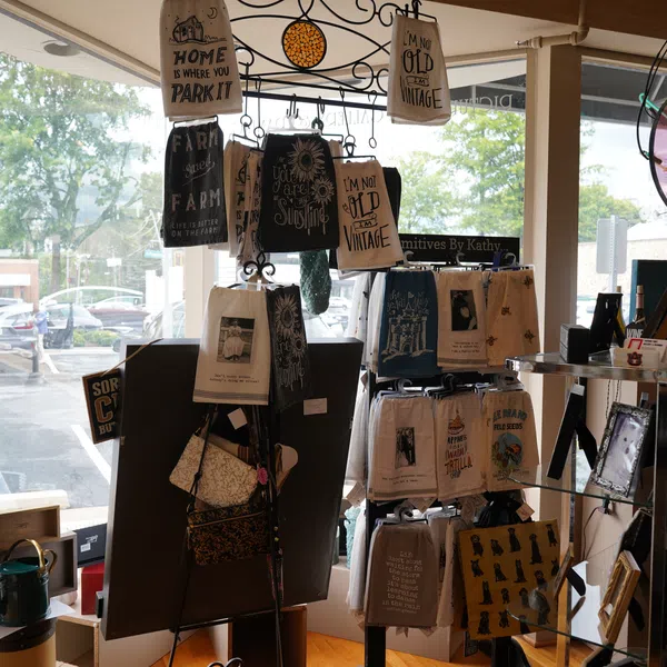 Store display with vintage-themed kitchen towels, signs, and framed prints near a large window with outdoor view.